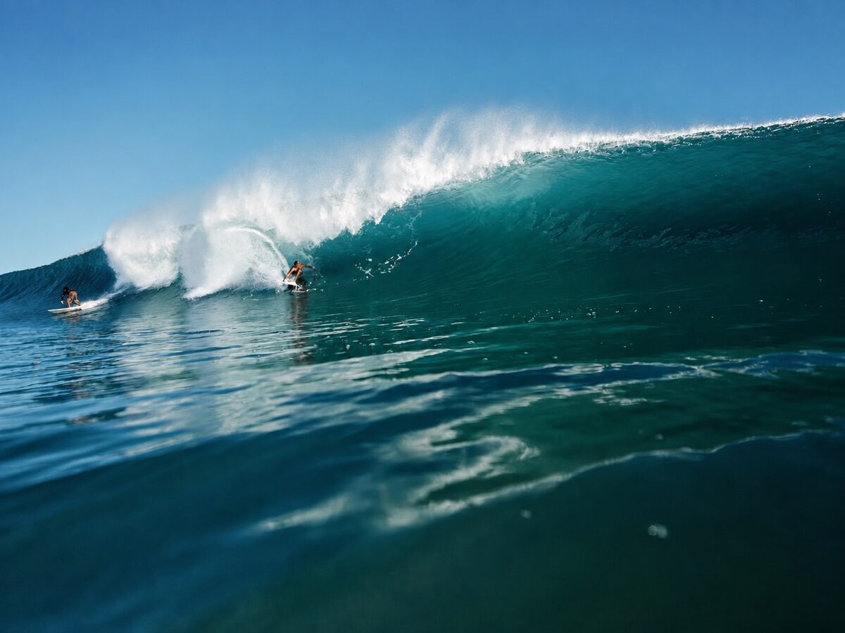 Two surfers riding a large breaking ocean wave under a clear blue sky