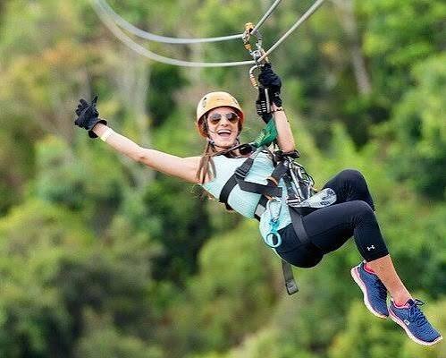 A smiling rider in a helmet and harness gliding along a zipline cable above green treetops